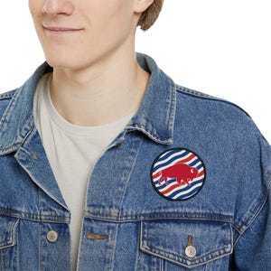 May include: A round patch featuring a red bison silhouette on a background of blue and white stripes and red stripes. The patch is sewn onto a denim jacket.