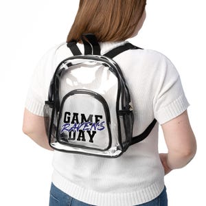 May include: Clear backpack with black trim and mesh side pockets. The front of the backpack features the text "GAME RAVENS DAY" in black and purple. The backpack has black adjustable straps and a top handle.