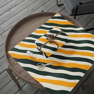 May include: A wrapped gift box with a brown ribbon and bow sits on striped wrapping paper. The paper features alternating bands of dark green, yellow, and off-white. A pair of scissors and tape are also on the paper, ready for wrapping.