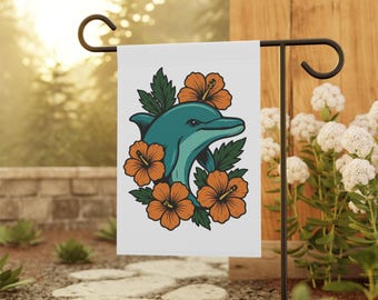 Puede incluir: Una bandera de jardín blanca con un delfín turquesa rodeado de flores de hibisco naranja y hojas verdes. La bandera se muestra en un soporte de metal negro en un jardín.