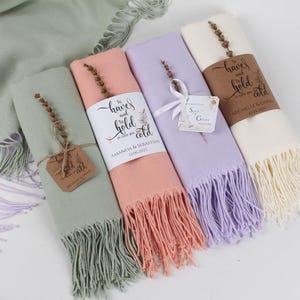 May include: Four pastel-coloured scarves, folded and presented. The colours are light green, peach, lilac, and cream. Each has a fringed edge and is wrapped with a band bearing text and a sprig of dried flowers. Tags with names and dates are attached.