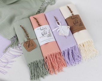 May include: Four pastel-coloured scarves, folded and presented. The colours are light green, peach, lilac, and cream. Each has a fringed edge and is wrapped with a band bearing text and a sprig of dried flowers. Tags with names and dates are attached.