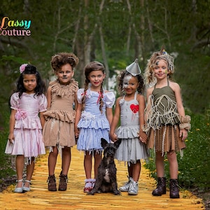 May include: Five children dressed as characters from The Wizard of Oz stand on a yellow brick road. Costumes include Dorothy, the Scarecrow, the Tin Man, and the Cowardly Lion. A small black dog sits in front of the children. The background is a forest.