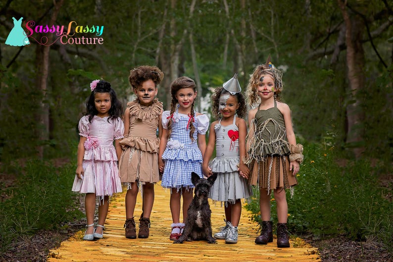 May include: Five children dressed as characters from The Wizard of Oz stand on a yellow brick road. Costumes include Dorothy, the Scarecrow, the Tin Man, and the Cowardly Lion. A small black dog sits in front of the children. The background is a forest.