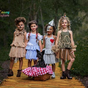 May include: Four children in Wizard of Oz costumes stand on a yellow brick road. Costumes include a lion, Dorothy, the Tin Man, and the Scarecrow. A black dog sits in a basket with a red and white checked cloth.