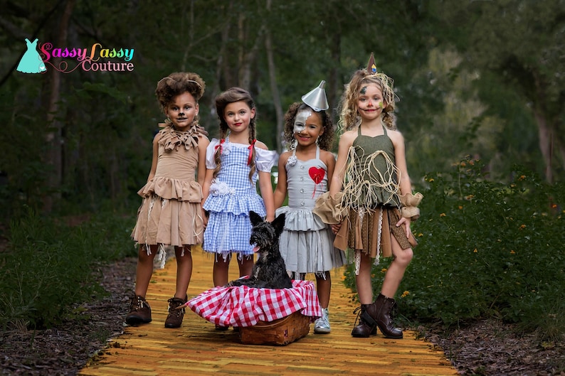 May include: Four children in Wizard of Oz costumes stand on a yellow brick road. Costumes include a lion, Dorothy, the Tin Man, and the Scarecrow. A black dog sits in a basket with a red and white checked cloth.