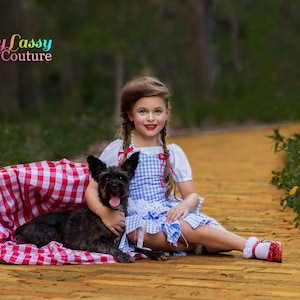 May include: A child in a blue and white gingham dress, white blouse, and red shoes, sitting on a yellow brick road with a black dog. A red and white checkered blanket is nearby. The image includes the text "Sassy Lassy Couture".