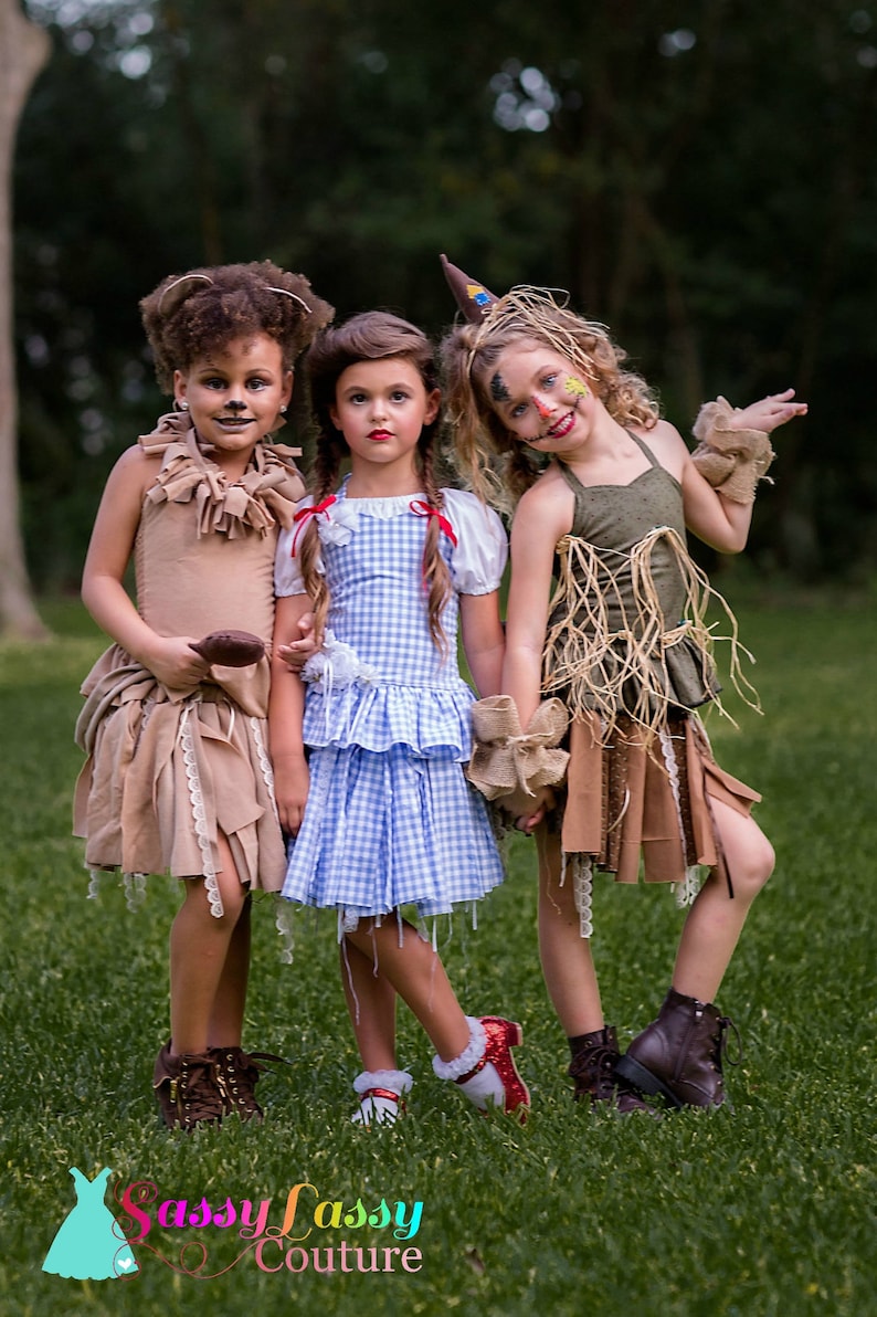 May include: Three children in Wizard of Oz costumes: a lion, Dorothy, and a scarecrow. The costumes are detailed and colourful, set against a green grassy background. The children are holding hands. The image is taken outdoors.