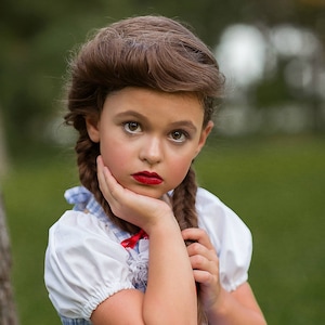 May include: A girl in a blue and white gingham dress with a white blouse and red bow. Her brown hair is styled in braids. She wears red lipstick and rests her hand on her cheek. The logo "Sassy Lassy Couture" is visible in the top right corner.