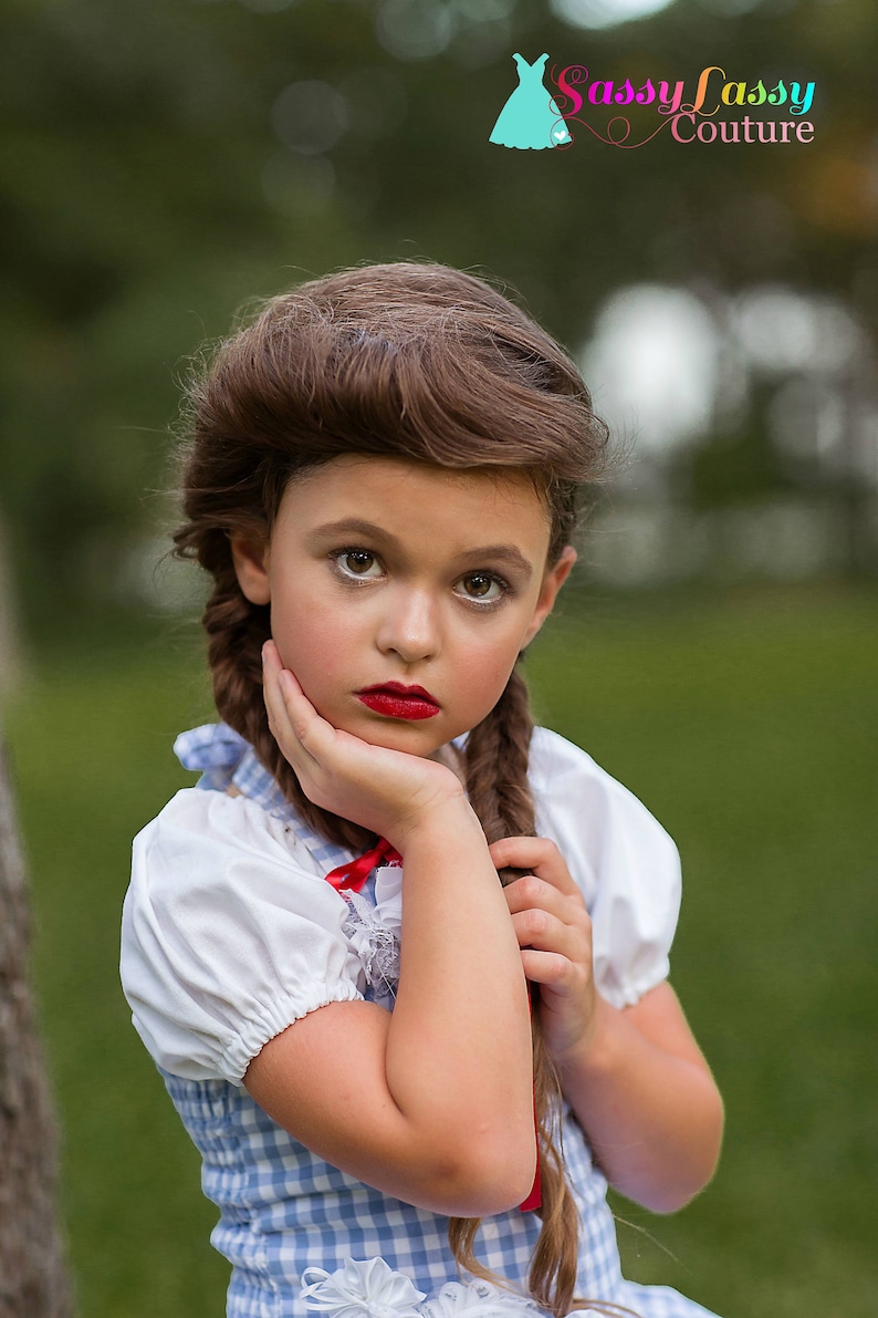 May include: A girl in a blue and white gingham dress with a white blouse and red bow. Her brown hair is styled in braids. She wears red lipstick and rests her hand on her cheek. The logo "Sassy Lassy Couture" is visible in the top right corner.