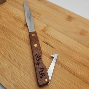 May include: A wooden handled stainless steel spreader with a small hook on the end. The handle is engraved with the words "Sling Pills".