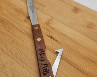 May include: A stainless steel and wood Sting Pills knife. The knife has a wooden handle with brass rivets and the words "Sting Pills" engraved on the side. The blade is open, revealing a second blade.