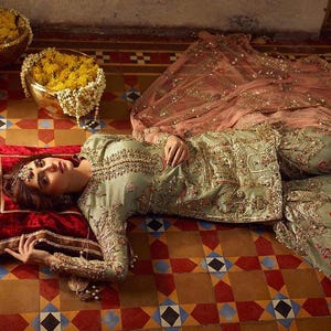 Peut inclure: Une femme portant une tenue brod&eacute;e verte et dor&eacute;e avec un dupatta rose est allong&eacute;e sur un sol en carrelage &agrave; motifs. La tenue est orn&eacute;e de perles et d'embellissements complexes.