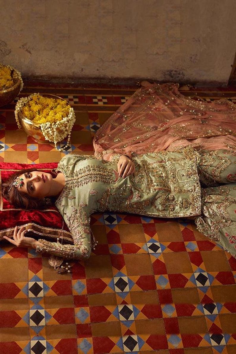 Peut inclure: Une femme portant une tenue brod&eacute;e verte et dor&eacute;e avec un dupatta rose est allong&eacute;e sur un sol en carrelage &agrave; motifs. La tenue est orn&eacute;e de perles et d'embellissements complexes.
