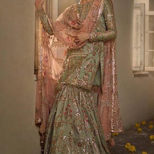Peut inclure: Une femme portant une robe brod&eacute;e verte et dor&eacute;e avec un dupatta rose. La robe a une longue jupe &agrave; bas &eacute;vas&eacute; et un corsage ajust&eacute;. Le dupatta est transparent et pr&eacute;sente un motif floral. La femme se tient dans une porte avec des fleurs jaunes sur le sol.