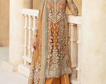 May include: A woman wearing a long, golden-brown dress with intricate embroidery and a matching dupatta. The dress has a sheer overlay and is adorned with silver embellishments. The woman is standing in front of a white pillar.