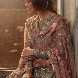 Peut inclure: Une femme portant une robe brod&eacute;e verte et rose avec un dupatta assorti. La robe est orn&eacute;e de perles et de paillettes. La femme est assise pr&egrave;s d'une fen&ecirc;tre.
