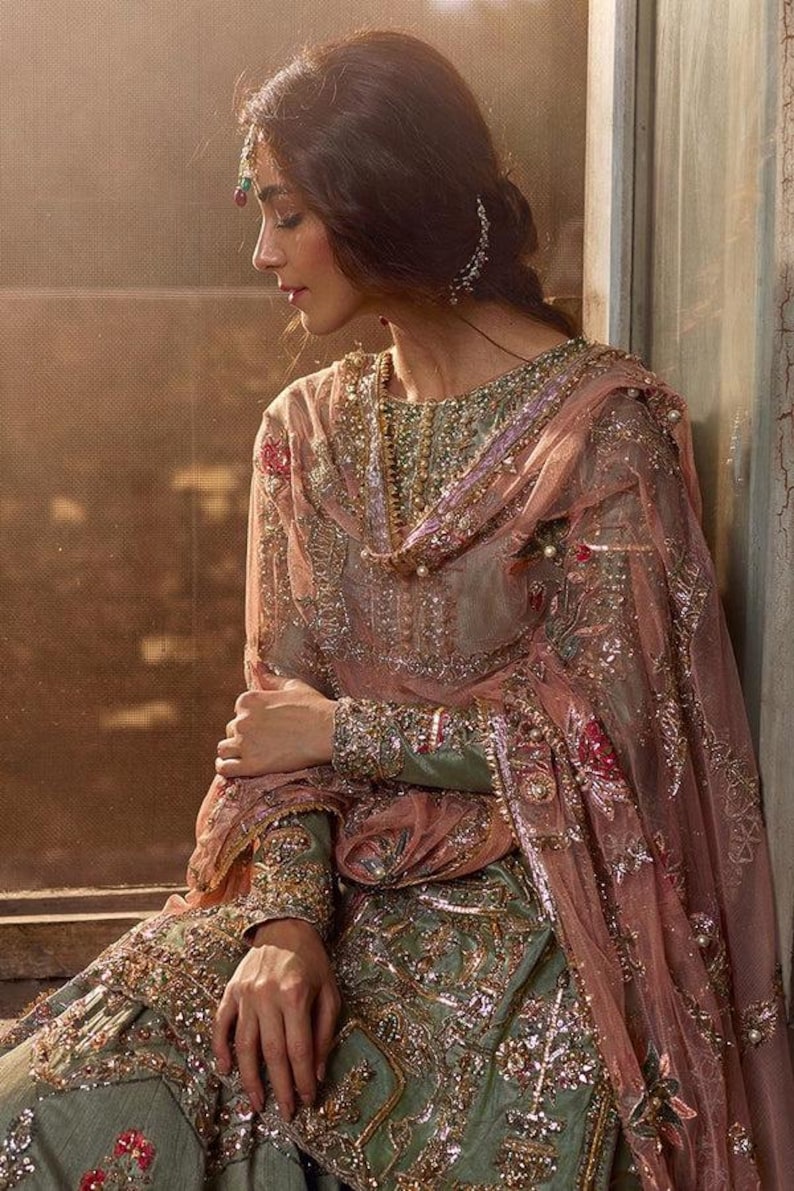 Peut inclure: Une femme portant une robe brod&eacute;e verte et rose avec un dupatta assorti. La robe est orn&eacute;e de perles et de paillettes. La femme est assise pr&egrave;s d'une fen&ecirc;tre.