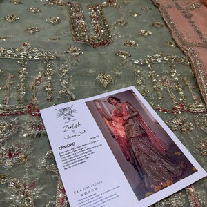 Peut inclure: Un tissu brod&eacute; vert et rose avec des embellissements dor&eacute;s. Le tissu est pr&eacute;sent&eacute; avec une &eacute;tiquette qui indique "Zarnish by Khushboo" et "Zamurd" avec une description du tissu. L'&eacute;tiquette comprend &eacute;galement des instructions d'entretien.
