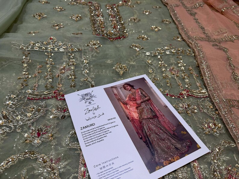 Peut inclure: Un tissu brod&eacute; vert et rose avec des embellissements dor&eacute;s. Le tissu est pr&eacute;sent&eacute; avec une &eacute;tiquette qui indique "Zarnish by Khushboo" et "Zamurd" avec une description du tissu. L'&eacute;tiquette comprend &eacute;galement des instructions d'entretien.