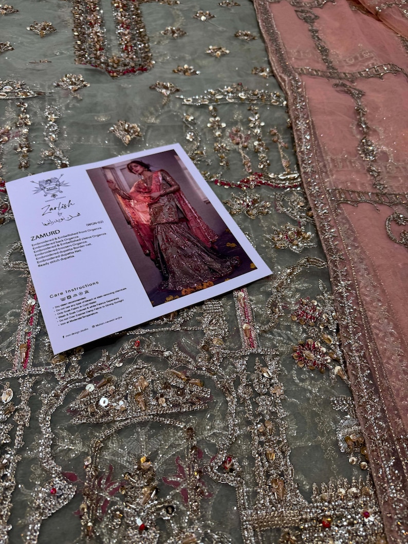 Peut inclure: Gros plan sur un tissu brod&eacute; vert et rose avec des paillettes et des perles. Le tissu est en filet et pr&eacute;sente un motif d&eacute;licat et complexe. Le tissu est pr&eacute;sent&eacute; avec une &eacute;tiquette qui indique "Zariish by Humaira" et "Zamurd".