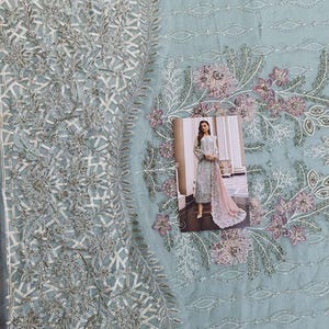 May include: A light blue embroidered fabric with a floral design. A woman in a light blue dress with a pink dupatta is standing in front of a window. The fabric is perfect for making a dress or other garment.