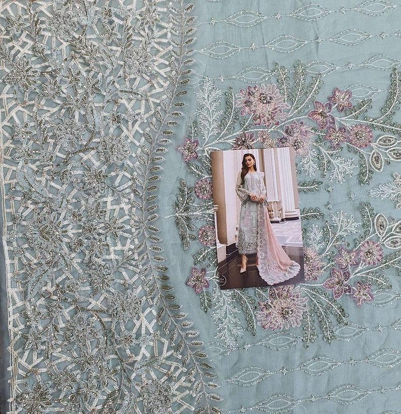 May include: A light blue embroidered fabric with a floral design. A woman in a light blue dress with a pink dupatta is standing in front of a window. The fabric is perfect for making a dress or other garment.