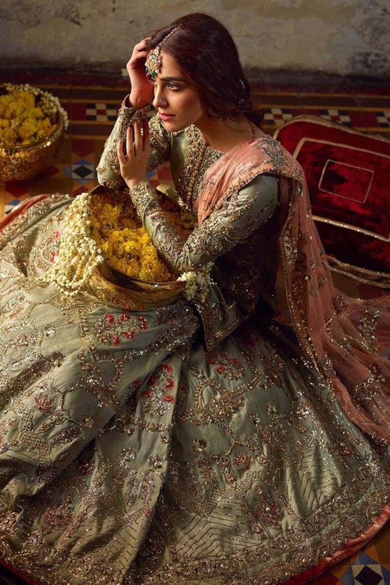 Peut inclure: Une femme portant une robe brod&eacute;e verte et dor&eacute;e avec un dupatta rose. Elle tient un bol de fleurs jaunes et est assise sur un sol &agrave; motifs.