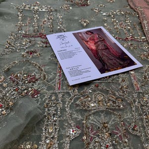 Peut inclure: Un tissu bleu clair avec des broderies argent&eacute;es et rouges complexes. Le tissu est d&eacute;cor&eacute; d'un motif r&eacute;p&eacute;titif d'arches et de motifs floraux. Une carte avec le texte "Zariyat by Wajahat Khan" est plac&eacute;e sur le tissu.