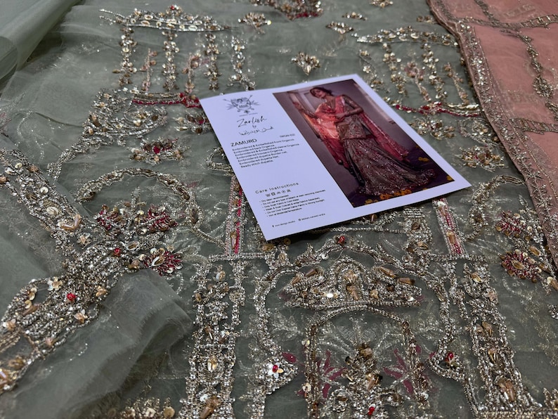 Peut inclure: Un tissu bleu clair avec des broderies argent&eacute;es et rouges complexes. Le tissu est d&eacute;cor&eacute; d'un motif r&eacute;p&eacute;titif d'arches et de motifs floraux. Une carte avec le texte "Zariyat by Wajahat Khan" est plac&eacute;e sur le tissu.