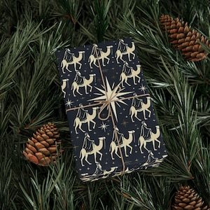 May include: A wrapped gift featuring a dark blue background with a repeating pattern of camels and riders in gold. The gift is tied with twine and a large star design. The background includes pine needles and pine cones.