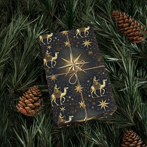 May include: Wrapped gift with a dark blue background and gold illustrations of camels, stars, and a large starburst. The gift is tied with twine and rests on a bed of green pine needles and pine cones, creating a festive holiday theme.