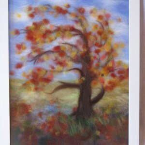 May include: A framed felt art piece depicting a tree in autumn. The tree's trunk is brown, with branches reaching out. The leaves are a mix of red, orange, and yellow. The background is a soft blue sky with hints of white.