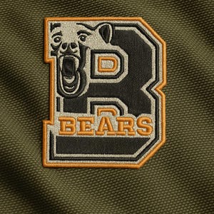 Könnte beinhalten: Gestickter Aufnäher mit einem stilisierten Bärenkopf in einem Buchstaben "B". Der Aufnäher hat einen schwarzen Hintergrund mit orangefarbenen und weißen Akzenten, und das Wort "BEARS" steht unten. Auf einem strukturierten olivgrünen Stoff.