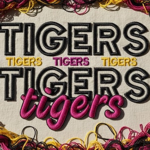 May include: Embroidered artwork featuring the word "TIGERS" in black outlined letters, with the word "TIGERS" in yellow and pink. The artwork is framed by a border of yellow, pink, and black thread.