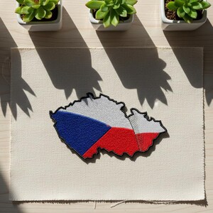 May include: Embroidered patch in the shape of the Czech Republic, featuring the national flag's colors: blue, white, and red. The patch is placed on a beige fabric, with three small potted succulents in the background.