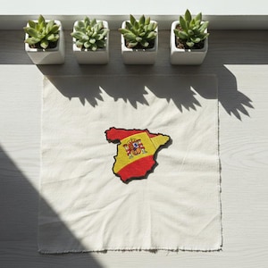 May include: A white, square fabric patch with a red, yellow, and black embroidered map of Spain. Four small, square white planters with green succulents are arranged above the patch on a white surface.