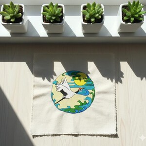 Può includere: Una toppa quadrata in tessuto bianco sporco con un disegno ricamato di una gru in volo contro un sole giallo e acqua blu. La toppa è esposta con quattro piccoli vasi quadrati bianchi, ciascuno con una pianta succulenta verde.
