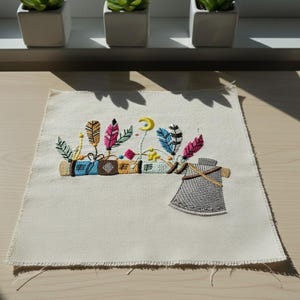 Puede incluir: Cuadrado de tela bordada con un diseño de hacha decorativa. El hacha es gris con un mango marrón, y la parte superior tiene plumas coloridas, una luna creciente y otros elementos decorativos. La tela es blanquecina.