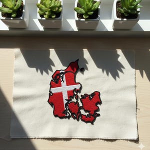 May include: A white fabric patch featuring an embroidered map of Denmark in red with a white cross. The patch is on a light-colored surface, with four small white square pots with green succulents in the background.