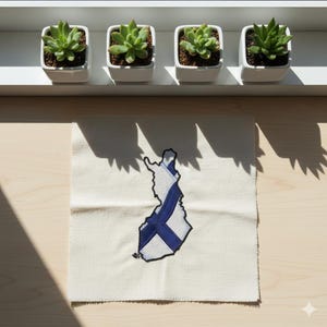 May include: A square, off-white fabric patch featuring an embroidered outline of Finland with a blue and white flag design. Four small, square white pots with green succulents sit on a white shelf above the patch. The image is lit by sunlight.