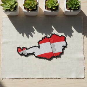 May include: Embroidered patch in the shape of Austria, featuring the Austrian flag's red, white, and red stripes. The patch is on a cream-colored fabric, with four small potted succulents above. The patch is approximately 3 inches long.