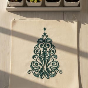 Puede incluir: Una funda de cojín cuadrada de color blanquecino con un diseño de árbol de Navidad bordado en verde azulado. El árbol presenta intrincados adornos y una estrella. Arriba, una bandeja blanca contiene cuatro pequeñas suculentas en macetas.