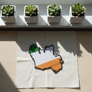 May include: Four small square white ceramic pots with succulents sit on a shelf. Below, a fabric patch displays the outline of Ireland, colored green, white, and orange, with a black border. The patch is on a cream-colored fabric.