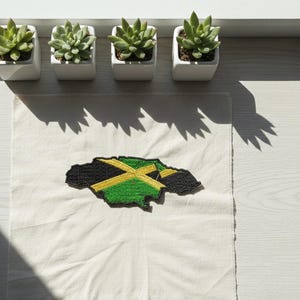 May include: An embroidered patch in the shape of Jamaica's island, featuring the country's flag colors: black, green, and yellow. The patch is placed on a white fabric, with four small potted succulents above.