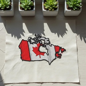May include: Embroidered patch featuring the map of Canada with the Canadian flag design. The patch is red and white with a black outline. The patch is on a cream-colored fabric. Four small potted succulents are in the background.