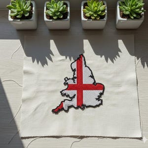 May include: Embroidered patch of England, with the St George's Cross in red on a white background. The patch is on a piece of off-white fabric. Four small potted succulents are in the background.