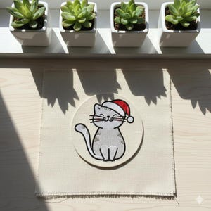 Peut inclure: Un écusson brodé représentant un chat gris portant un bonnet de Père Noël rouge et blanc, sur un carré de tissu blanc cassé. Quatre petites plantes succulentes dans des pots carrés blancs sont sur un rebord de fenêtre.