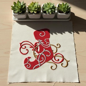 Puede incluir: Un calcetín navideño rojo con detalles bordados en blanco y dorado sobre tela color crema. El calcetín está decorado con patrones arremolinados. Cuatro pequeñas suculentas en macetas están encima de la tela.