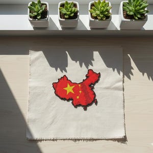 Op de afbeelding: Een rood-gele geborduurde patch in de vorm van de kaart van China, met een grote gele ster en kleinere sterren. De patch ligt op een vierkante, gebroken witte stof met een gerafelde rand. Vier kleine vetplanten in potten staan erboven.